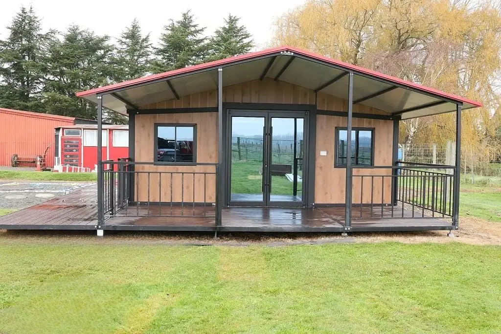 Front view of a 30ft expandable house featuring a covered deck, black-framed windows and doors, and a red gable roof, situated in a green rural setting.